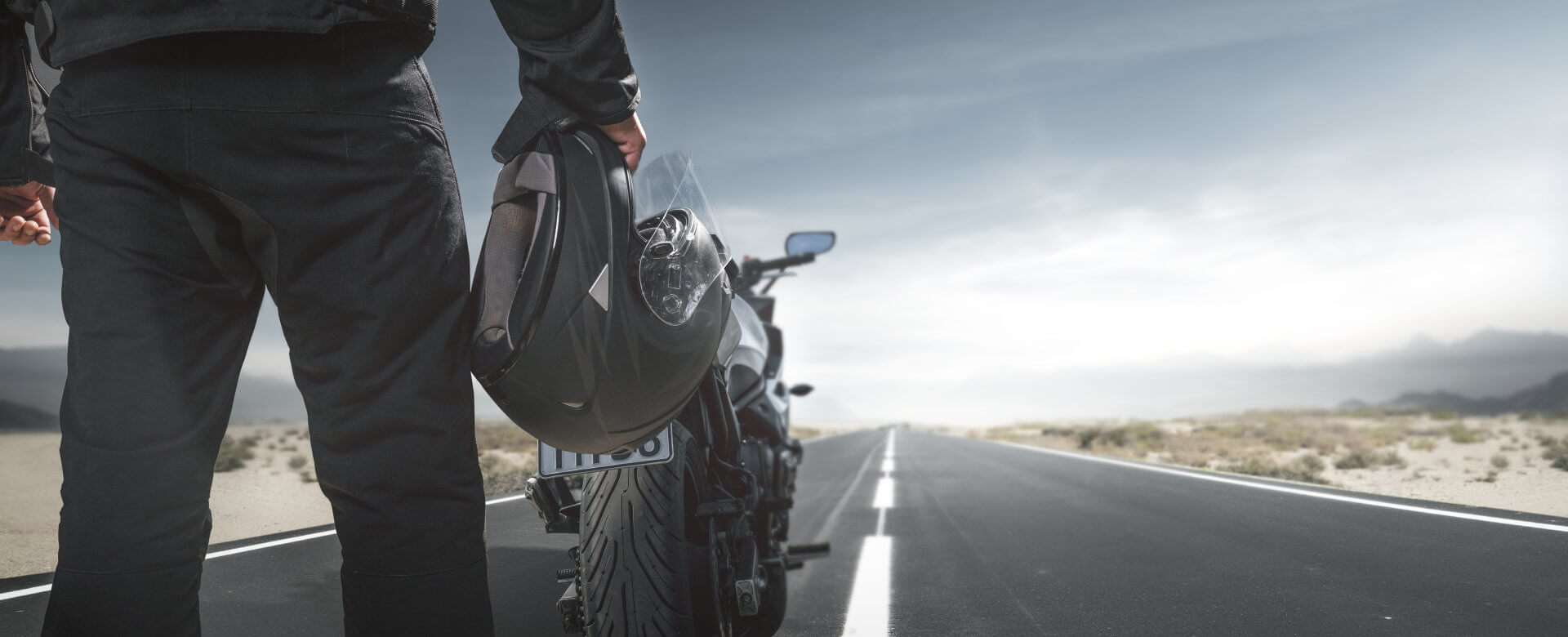 Ein Motorradfahrer hält seinen Helm in der Hand und steht mit seinem Motorrad auf einer Straße